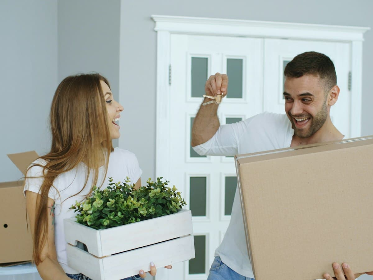A couple carrying boxes and a plant into their new home